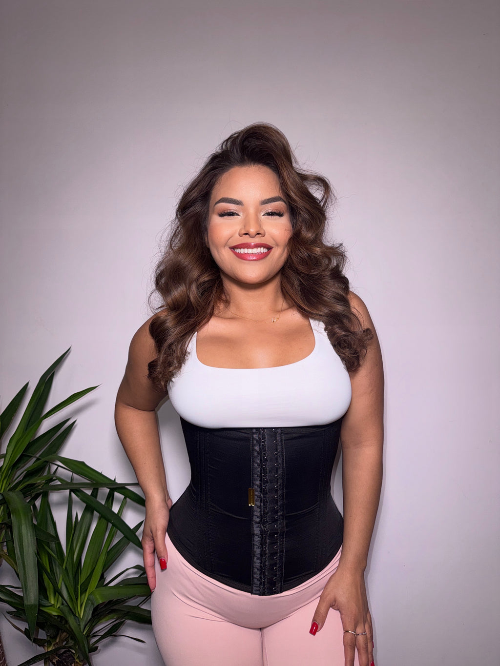 Woman wearing a black waist trainer with a white top and pink pants against a plain background.
